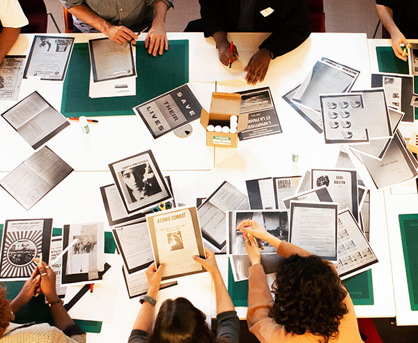 A group of people sit at an illuminated table covered in photocopied papers and materials