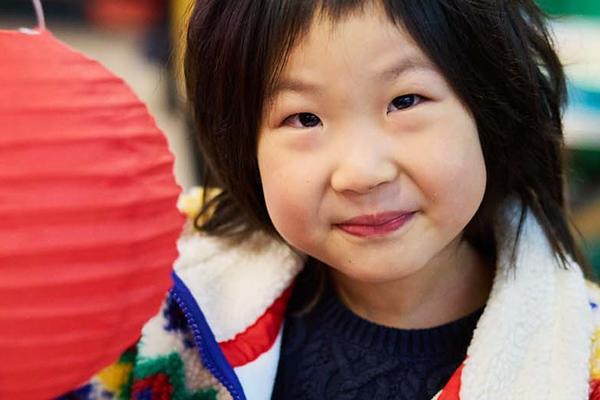 A girl with dark hair wearing a brightly coloured fleece holds up a red lantern 