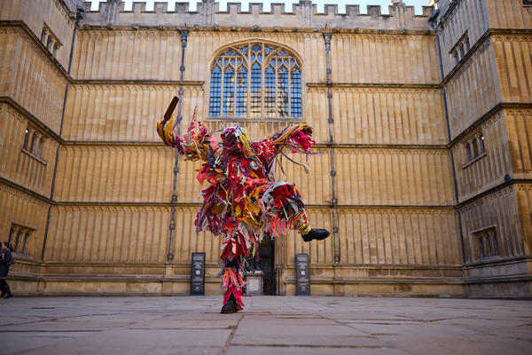 A person in a colourful costume made from strips of material dances in front of the entrance of the Bodleian Old Library
