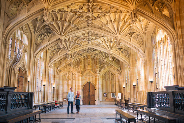 Two people stand in a large room with an ornately carved stone ceiling