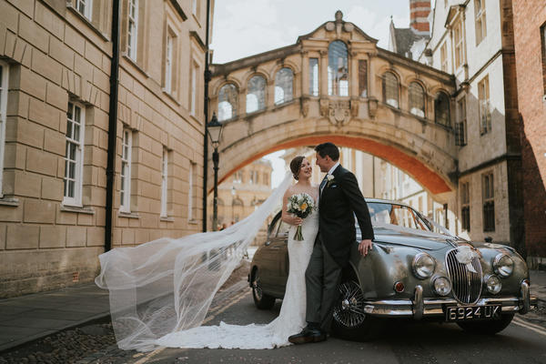 A couple in their bridal outfits stand in front of a vintage silver car underneath the Bridge of Sighs in Oxford